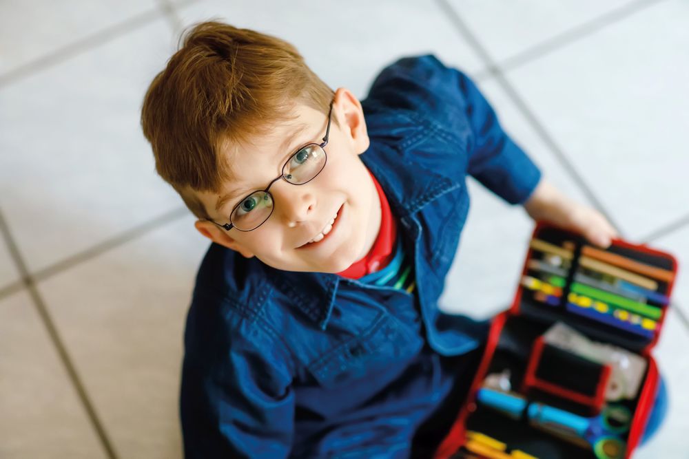 Happy little school kid boy searching for a pen in pencil case. Healthy schoolchild with glasses grab thinks for lessons in elementary class.